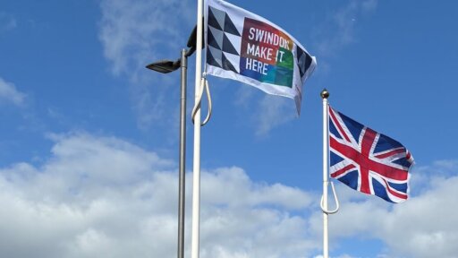 Two flags fly on tall flagpoles against a blue sky with scattered clouds. One flag features a geometric pattern with the words “Swindon Make It Here,” and the other is the United Kingdom flag.