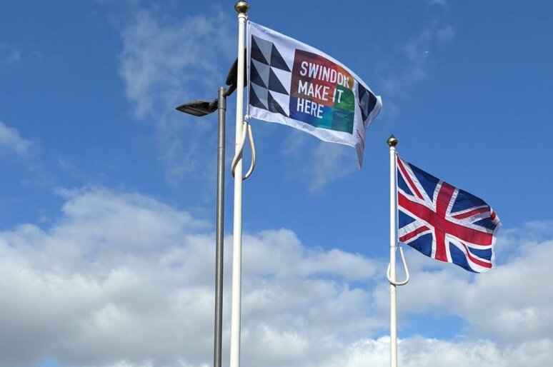 Two flags fly on tall flagpoles against a blue sky with scattered clouds. One flag features a geometric pattern with the words “Swindon Make It Here,” and the other is the United Kingdom flag.