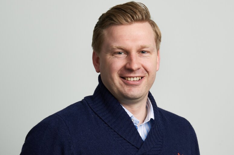 Ed Tellwright wearing a dark blue sweater with a small red embroidered logo and a light blue collared shirt underneath, posed against a plain light gray background in a clean, professional portrait style